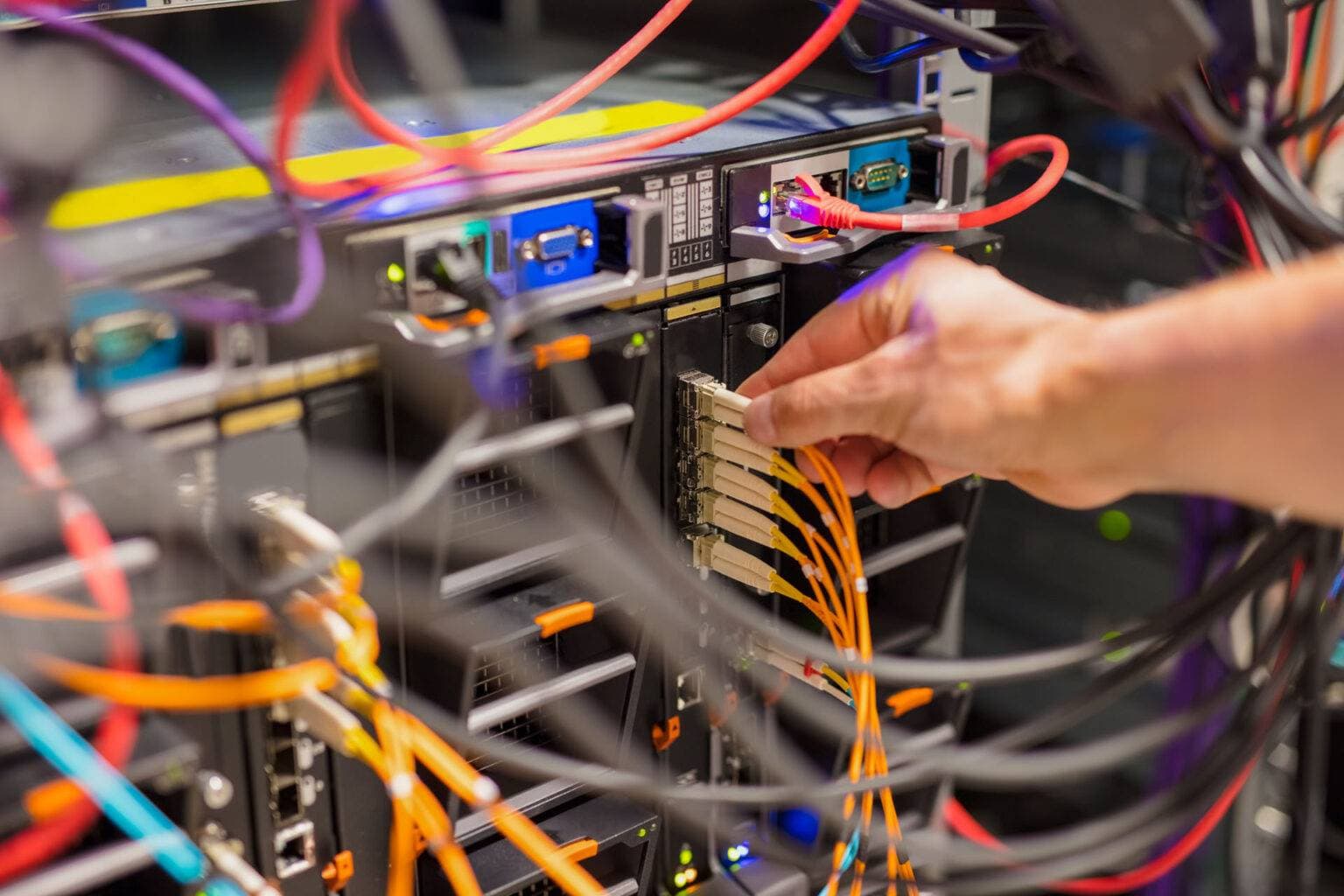 A hand connects yellow network cables to server rack ports amid various colored wires and electronic equipment, showcasing Resolute Technology LLC’s cabling expertise.