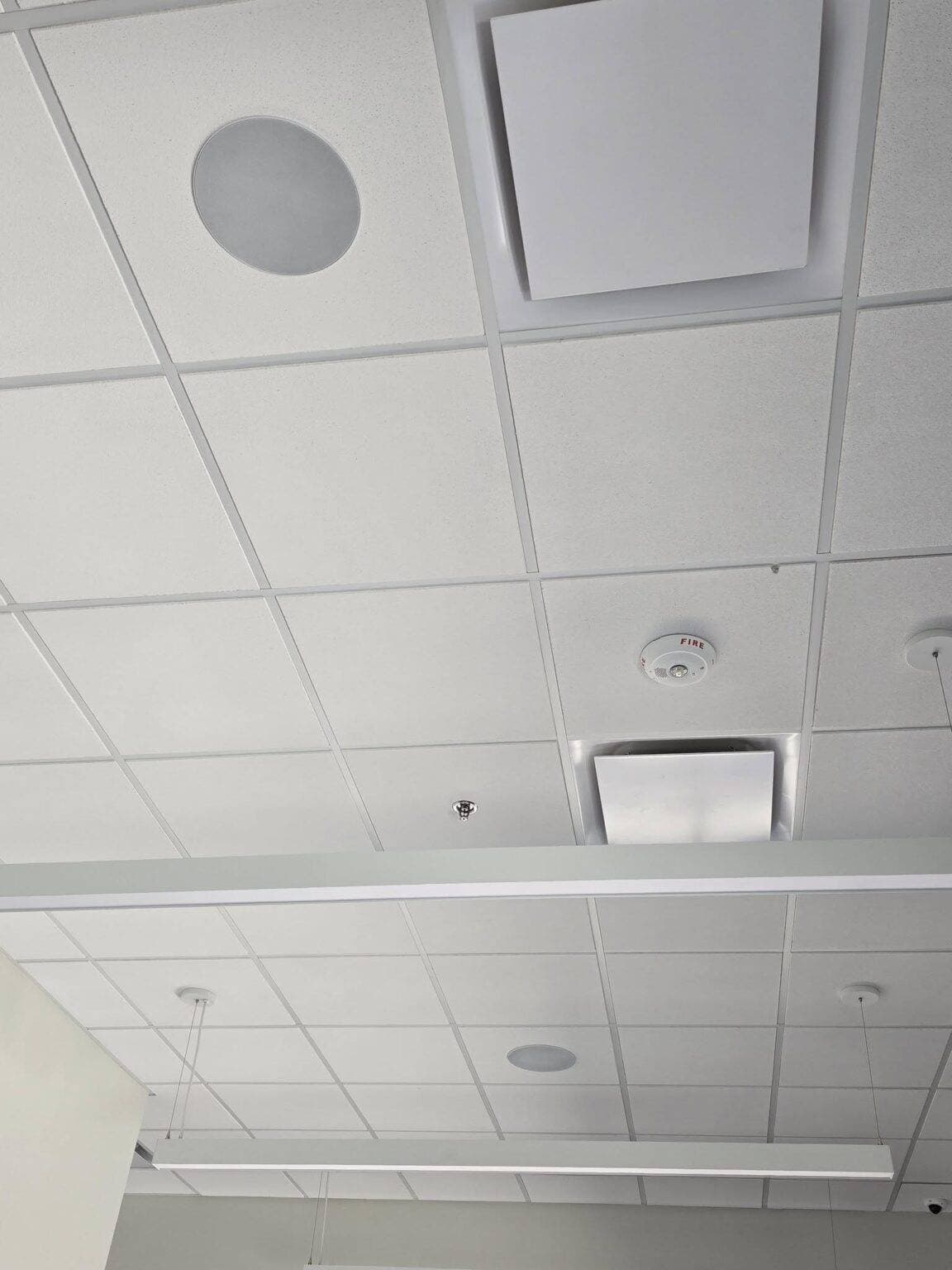 Commercial ceiling with white square panels, circular and square vents, a smoke detector, exposed wire, two hanging lights, and a structural beam.