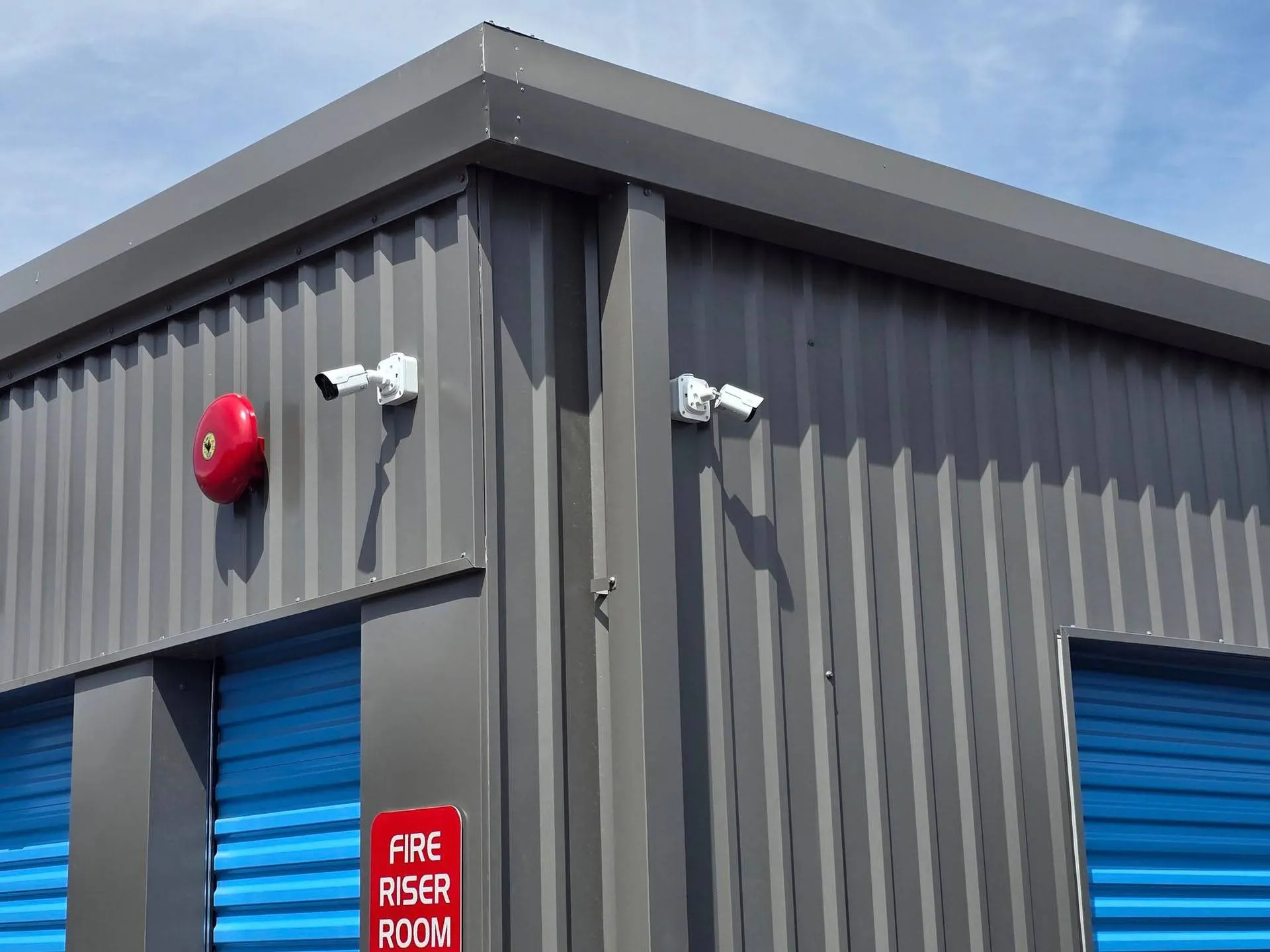 Gray metal building corner with cabling and two security cameras, a red fire alarm, a 