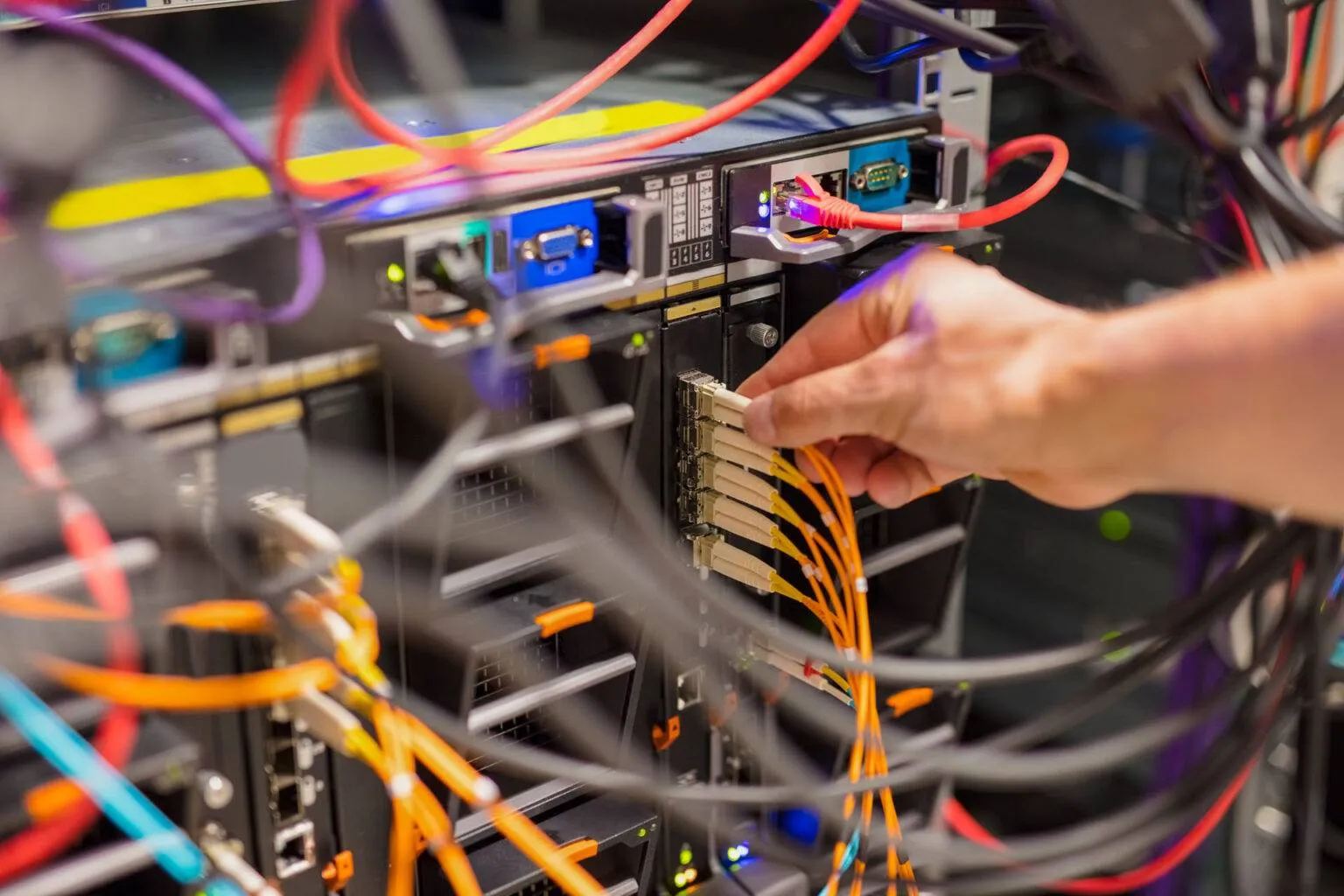 A hand connects yellow network cables to server rack ports amid various colored wires and electronic equipment, showcasing Resolute Technology LLC’s cabling expertise.