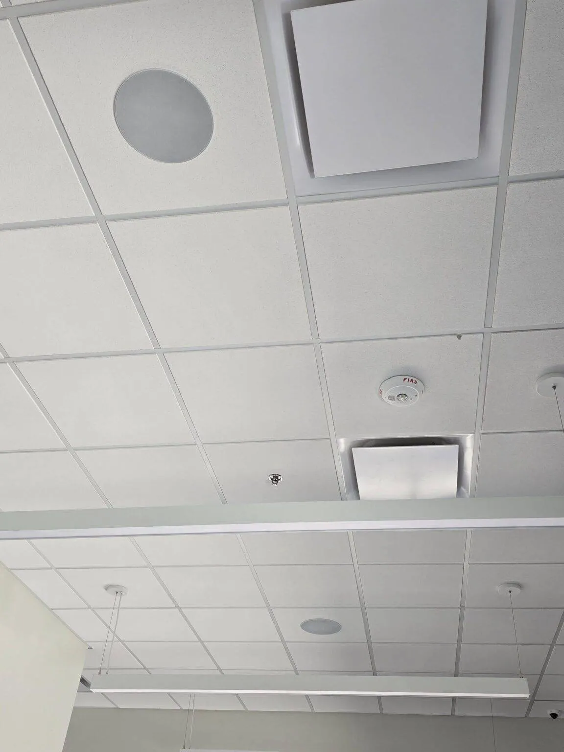 Commercial ceiling with white square panels, circular and square vents, a smoke detector, exposed wire, two hanging lights, and a structural beam.
