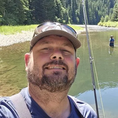 A licensed low voltage contractor takes a selfie while fishing in a river, with another person fishing in the background, surrounded by trees.