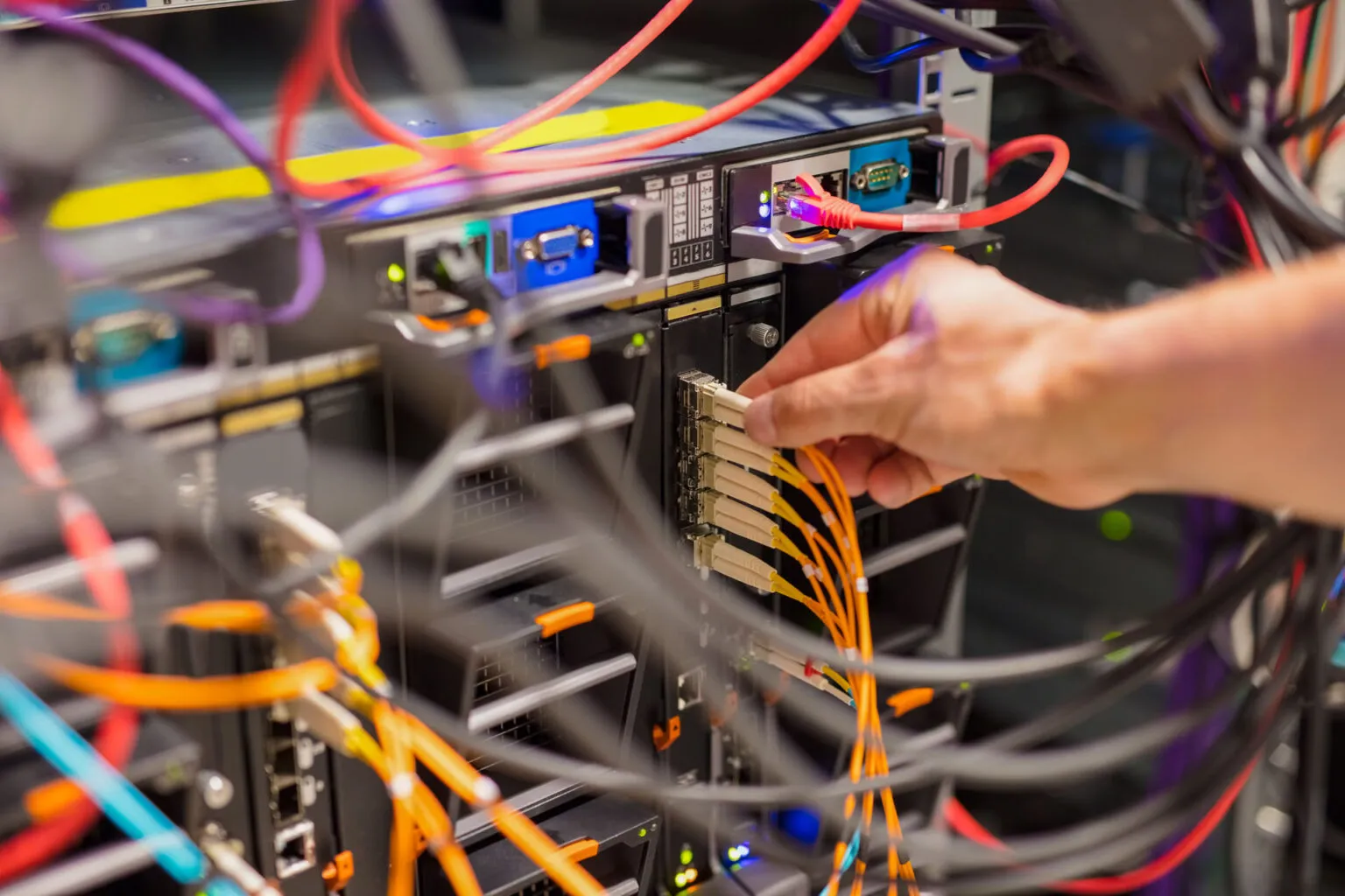 A hand connects yellow network cables to server rack ports amid various colored wires and electronic equipment, showcasing Resolute Technology LLC’s cabling expertise.
