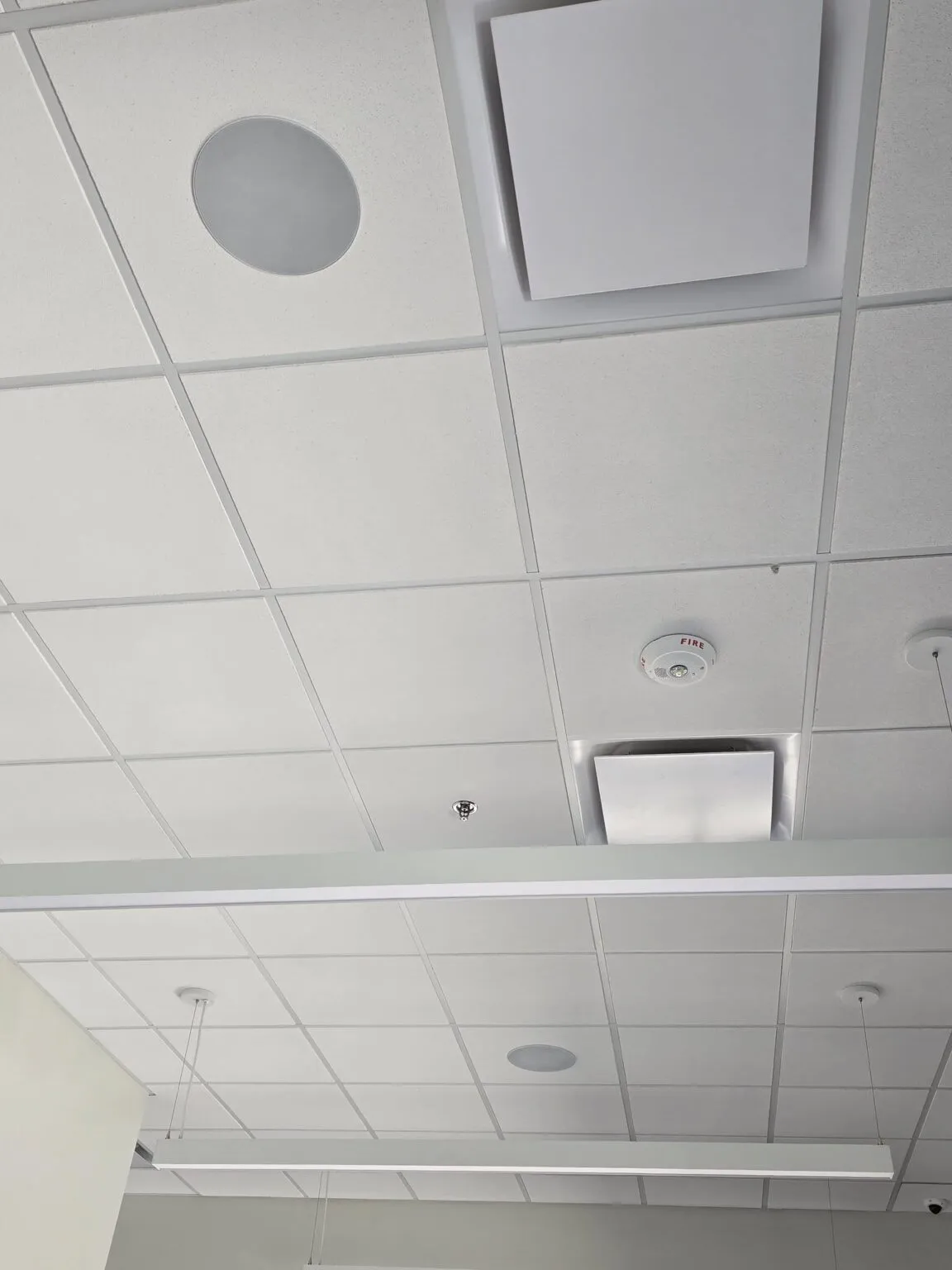 Commercial ceiling with white square panels, circular and square vents, a smoke detector, exposed wire, two hanging lights, and a structural beam.