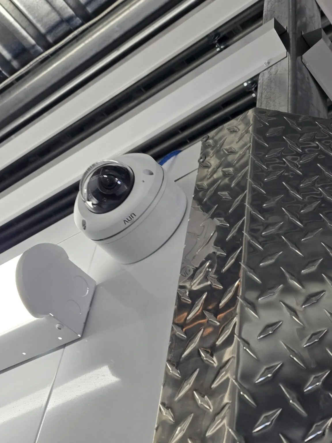 A dome security camera by Resolute Technology LLC is mounted on a wall near a metal surface with a diamond plate pattern in an industrial setting, providing reliable CCTV surveillance.