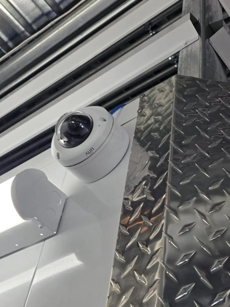 A dome security camera by Resolute Technology LLC is mounted on a wall near a metal surface with a diamond plate pattern in an industrial setting, providing reliable CCTV surveillance.