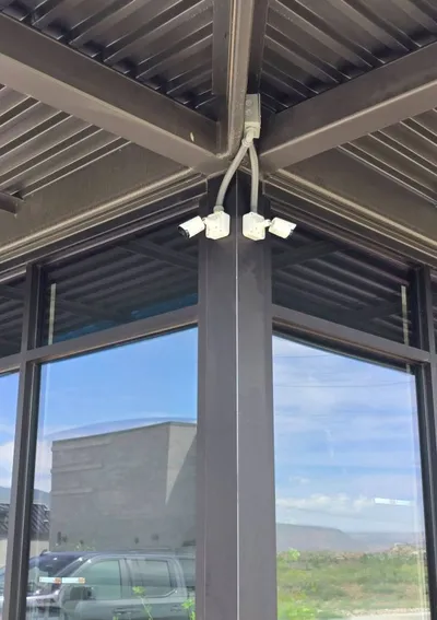 Four commercial security cameras are mounted under the metal roof at the corner of a building, with cabling neatly installed and all cameras aimed in different directions above glass windows.